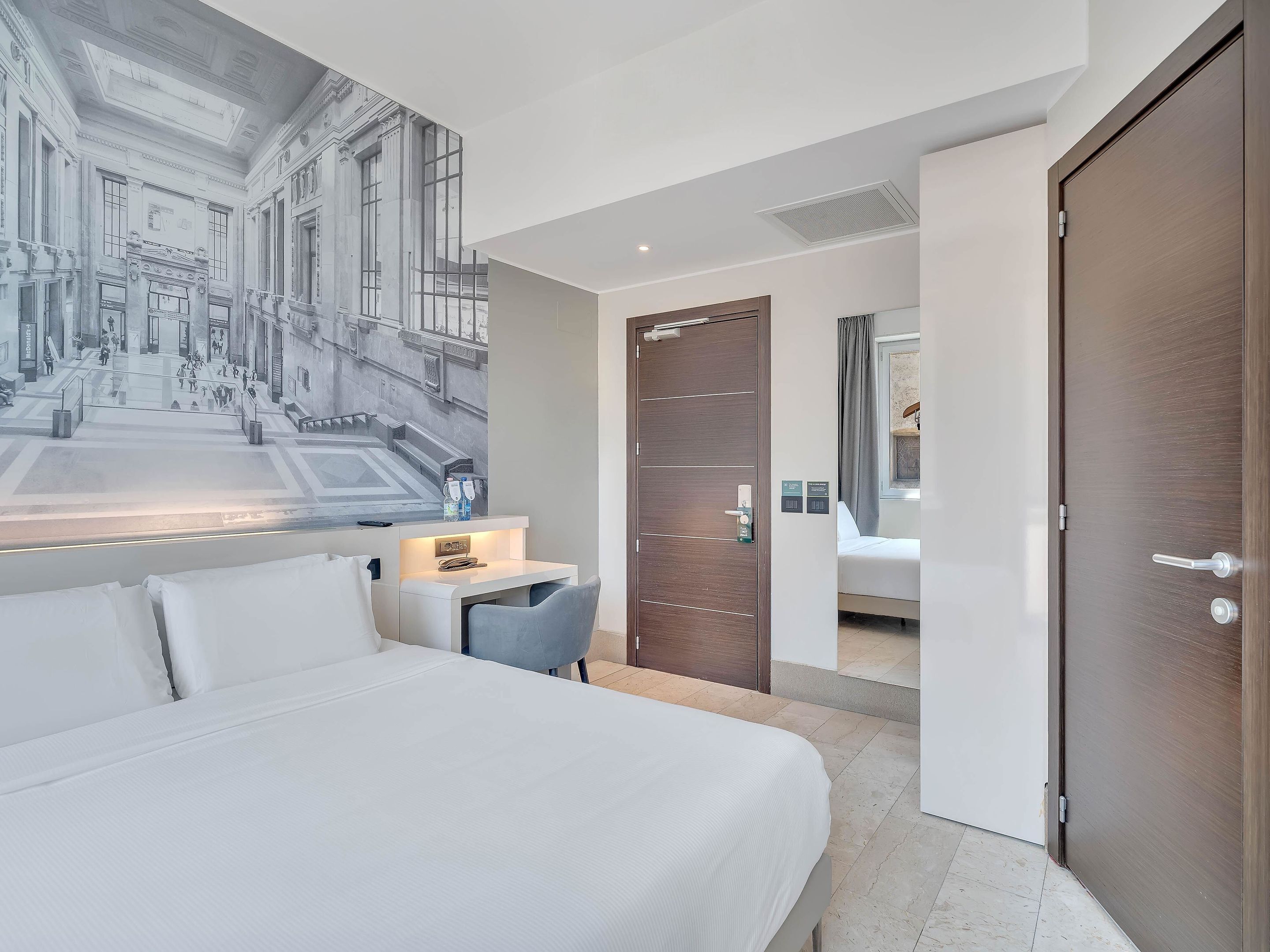 Una habitación de hotel con una cama grande, un escritorio con una silla, una puerta y una obra de arte colgada en la pared que representa un edificio.