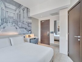 Hotelkamer met een groot bed, een bureau met stoel, een deur en een kunstwerk aan de muur met daarop een gebouw.
