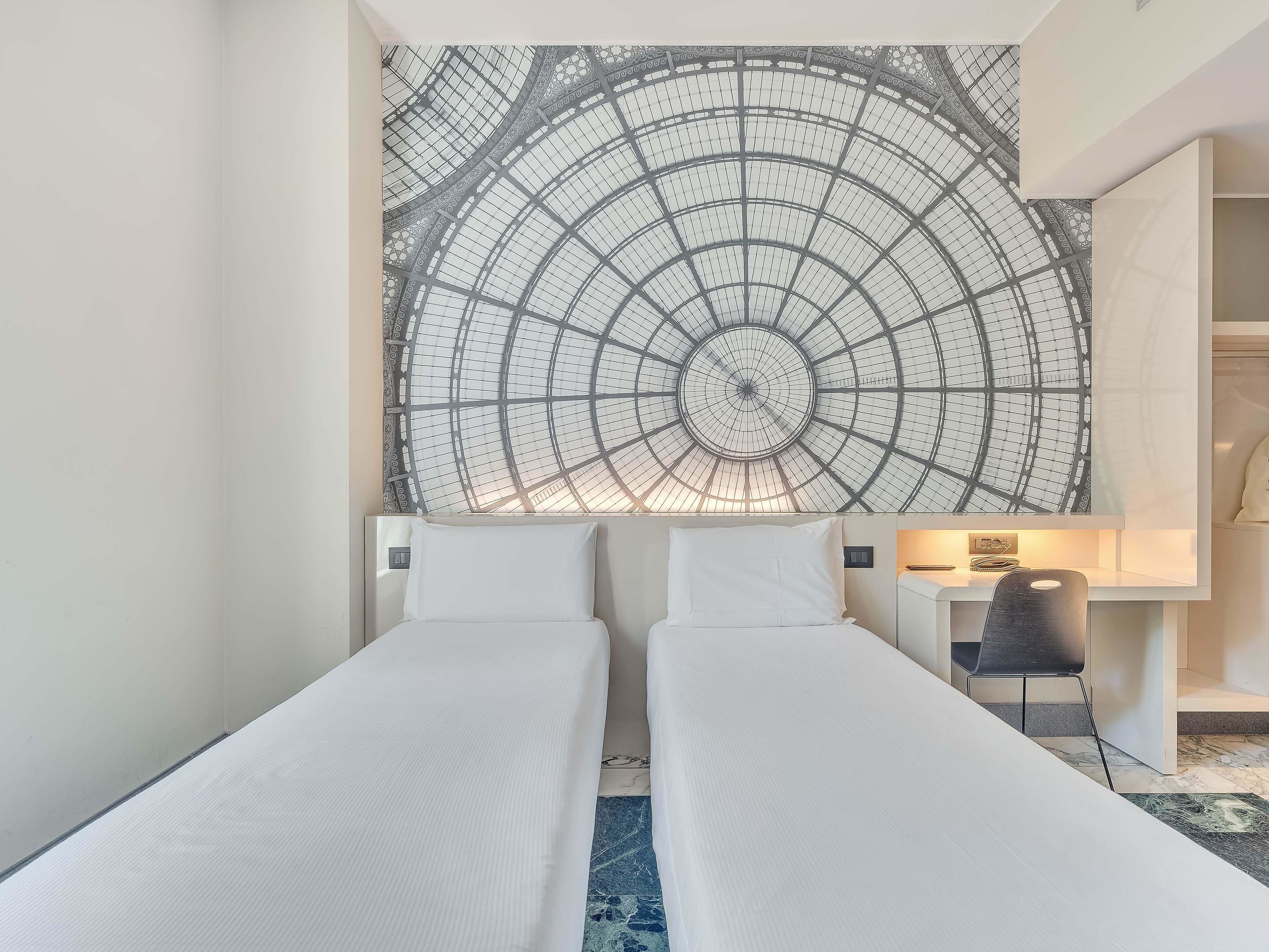 Hotellihuone, jossa on kaksi erillistä sänkyä, suuri seinävaate, työpöytä tuolilla ja yöpöytä.