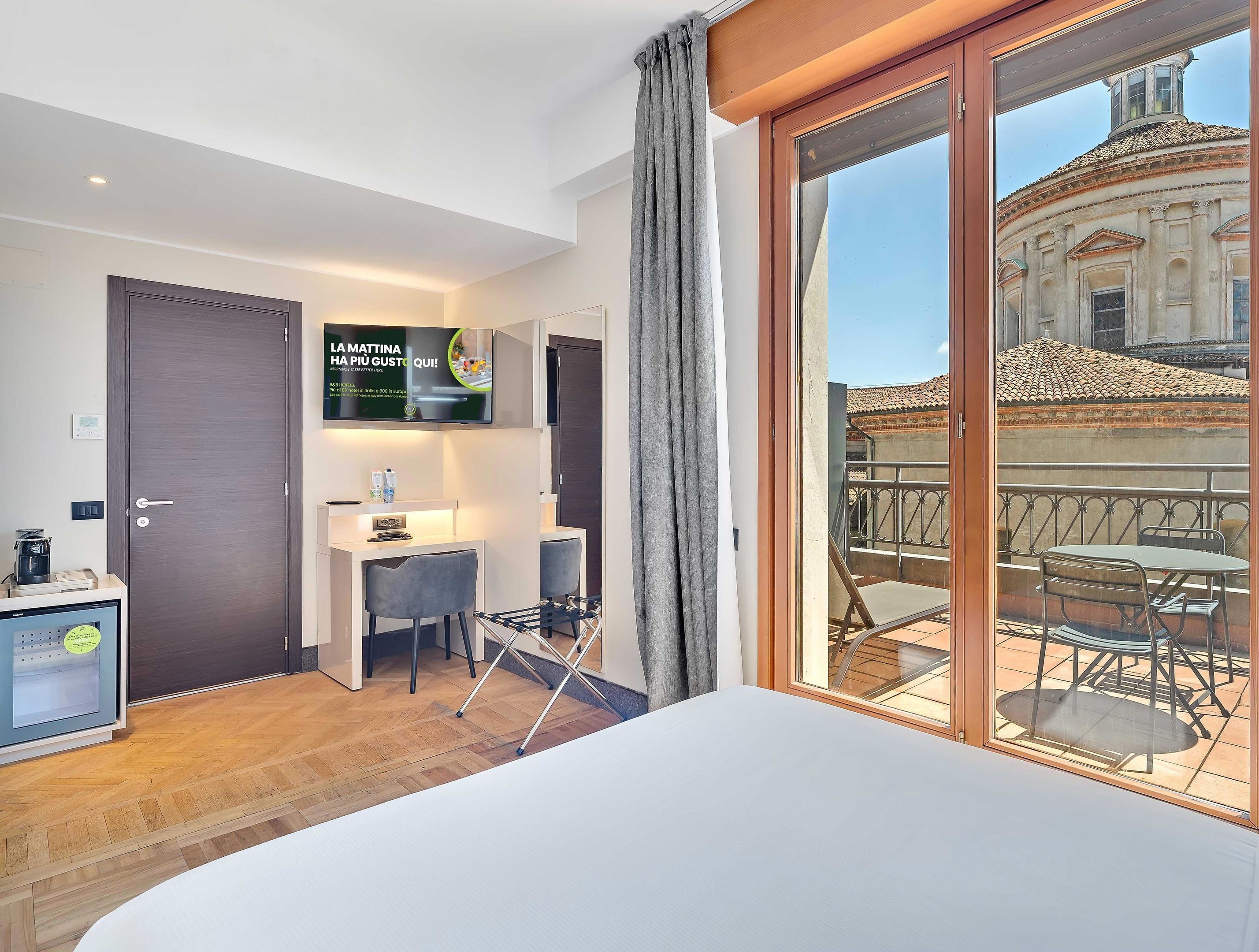 Habitación de hotel con cama, escritorio, silla, televisor y balcón con vista a un edificio.