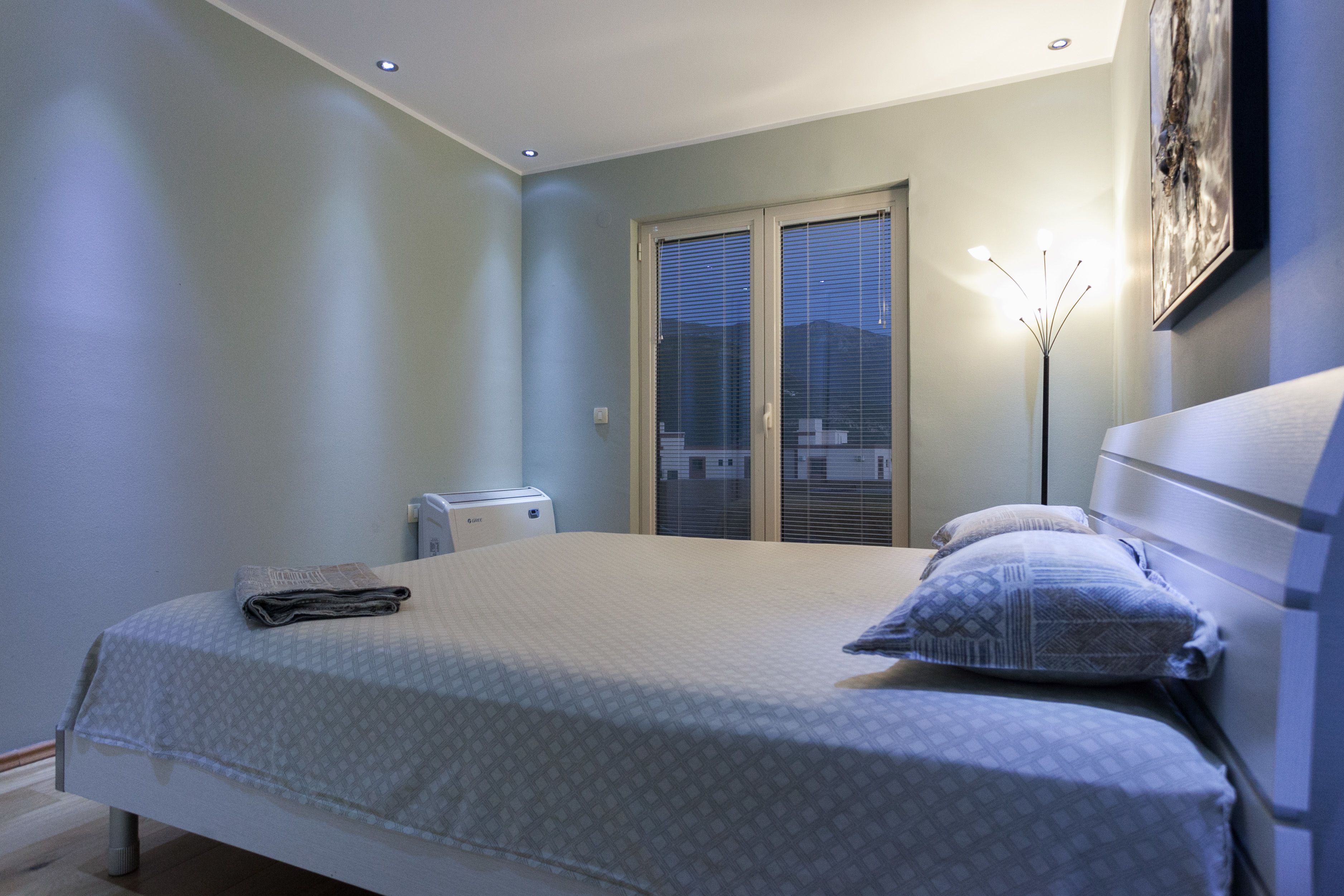 A bedroom with a bed, a bedside table, a lamp, and a window with blinds.