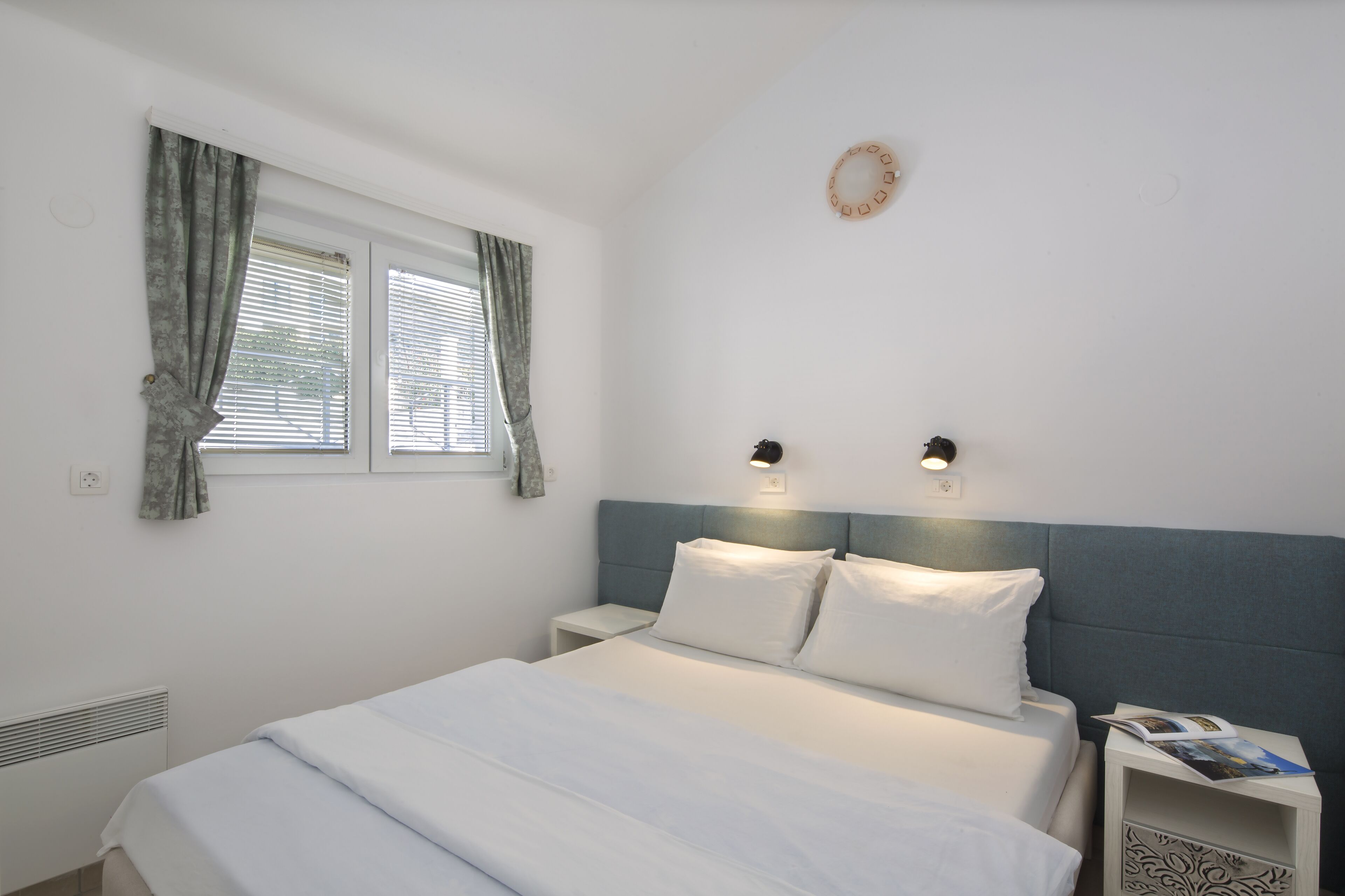A bedroom with a bed, two bedside tables, a window with curtains, and a wall-mounted light fixture.