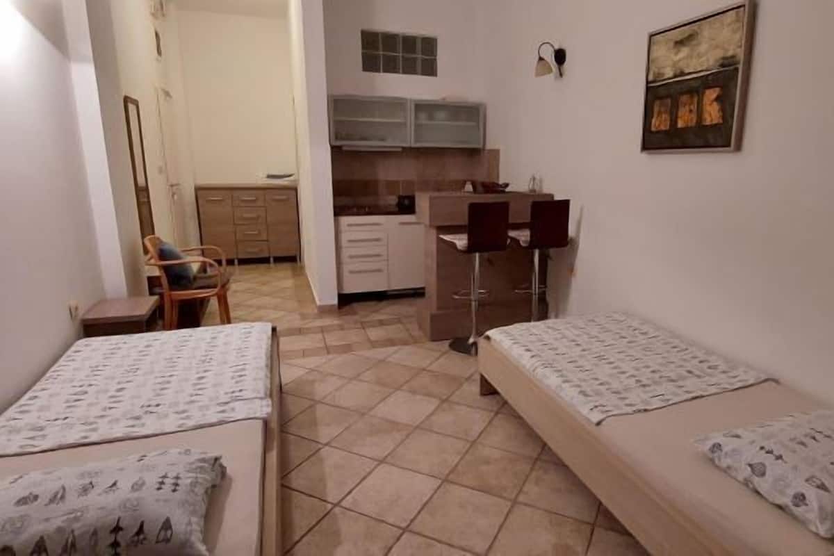 Una habitación pequeña y funcional con dos camas individuales, un kitchenette y una silla.