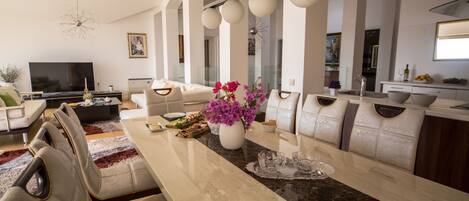 A modern living room with a large TV, comfortable seating, a dining area with a marble table, and a kitchen in the background.