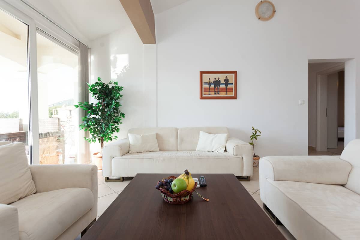 Una sala de estar moderna con un sofá blanco, una mesa de centro de madera con una cesta de frutas, y un ventanal que ofrece vistas al exterior.