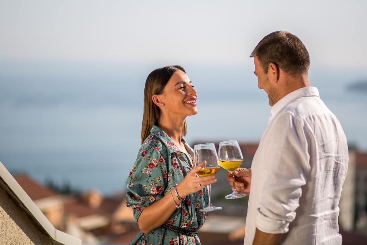 Una pareja sosteniendo copas de vino en un balcón con vistas a edificios y al mar.