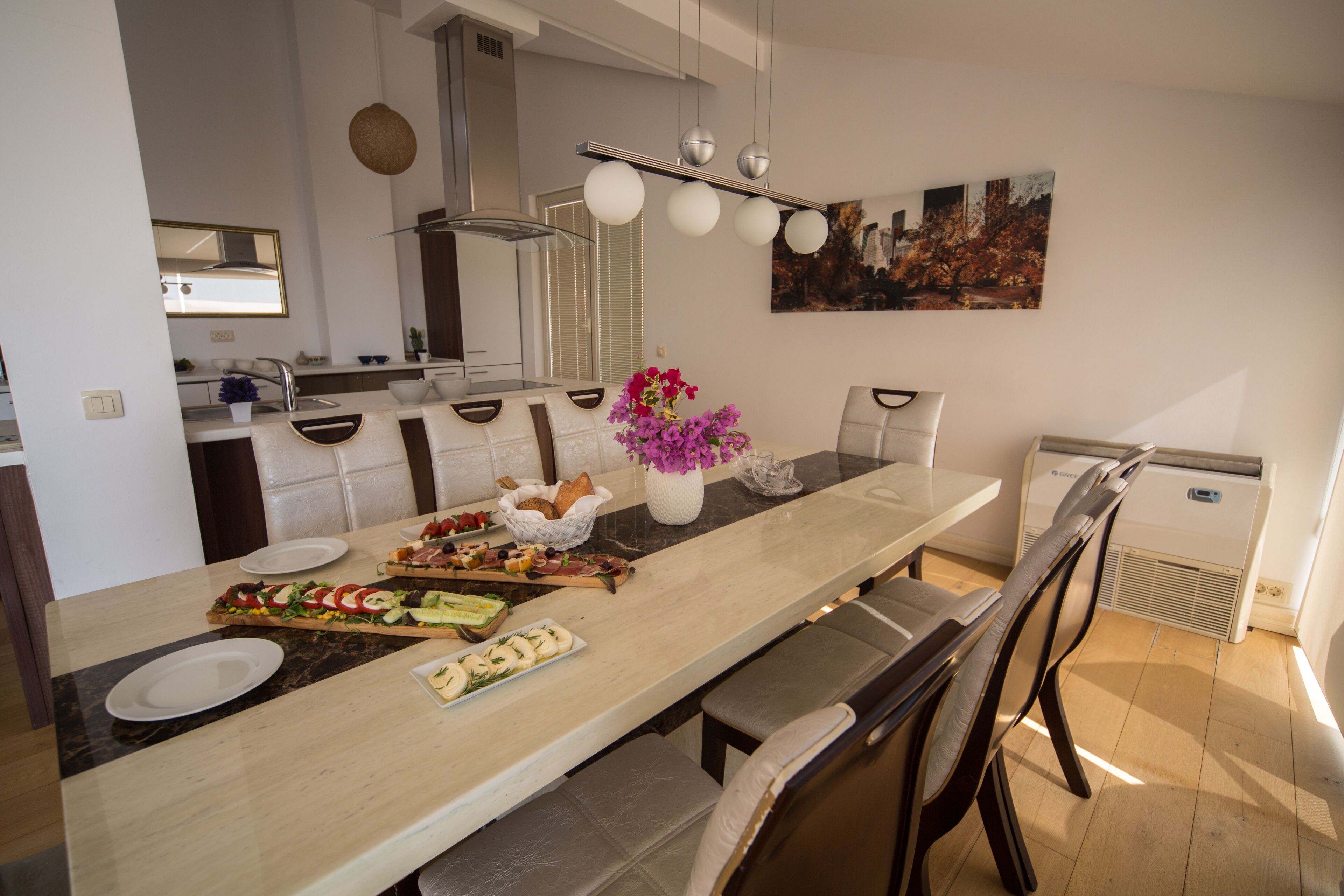 A modern kitchen with a long island, white countertops, and a central island with food. There are chairs around the island, a sink, and a stove.