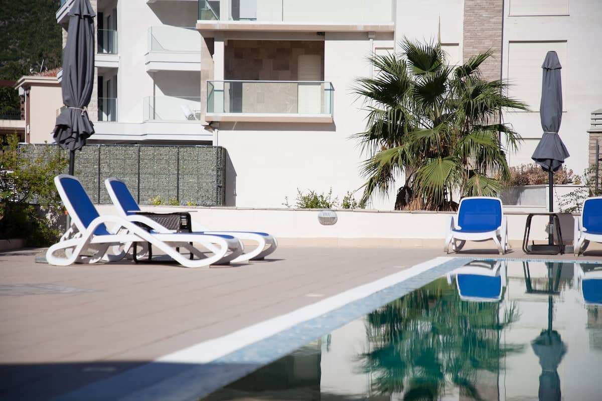 Zona de piscina con sillones de descanso y sombrillas, una palmera y un edificio moderno al fondo.