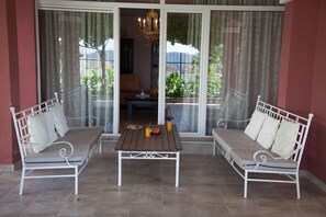 A balcony with two white metal sofas, a wooden table, and a glass door leading to a garden. - Spa Resort Becici (Becici)