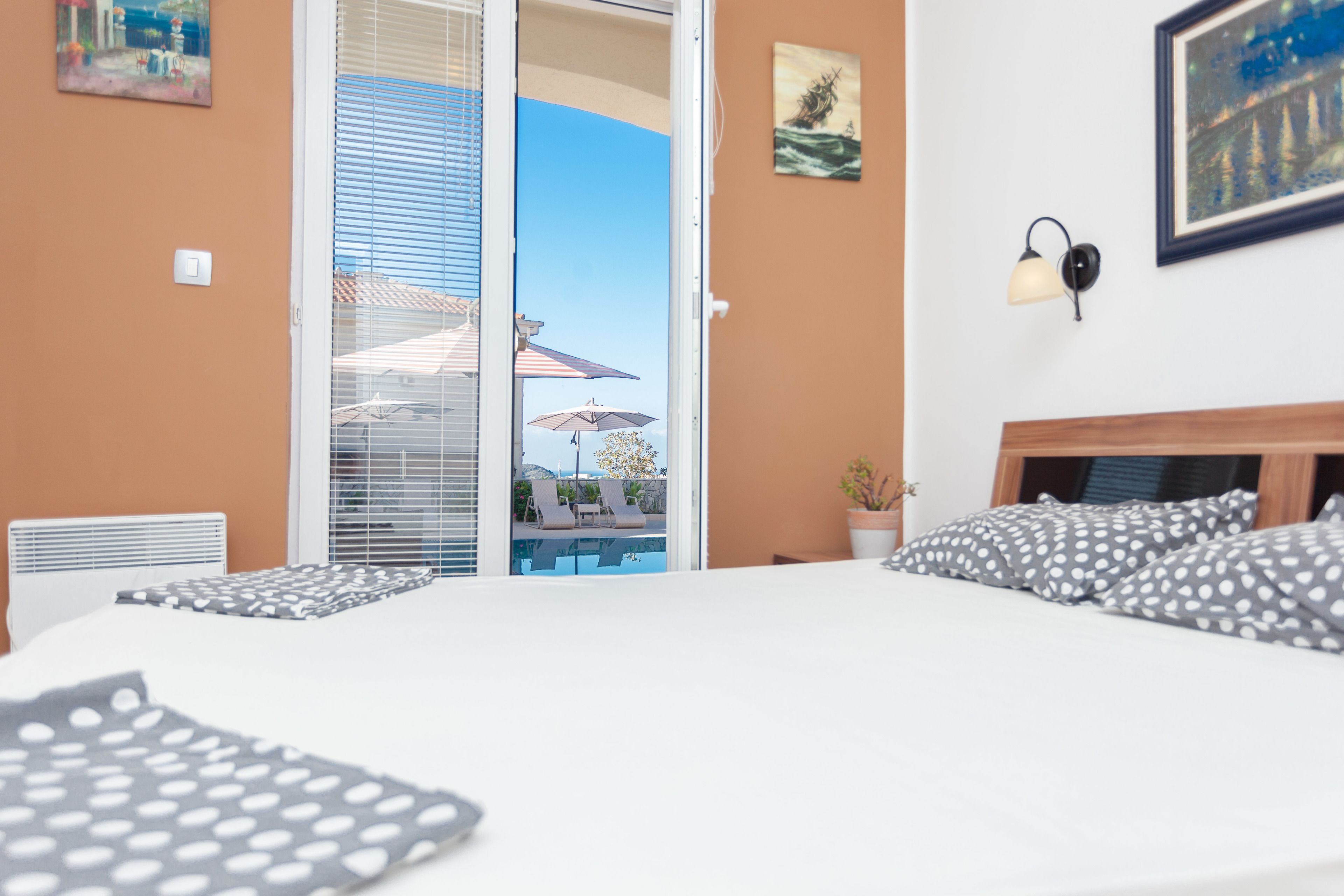 A bedroom with a bed, a nightstand, a potted plant, and a view of a pool and lounge chairs through a sliding door.