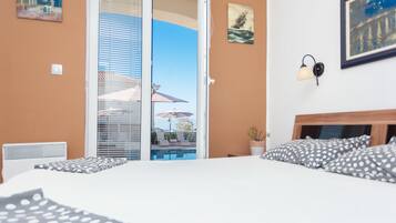 A bedroom with a bed, a nightstand, a potted plant, and a view of a pool and lounge chairs through a sliding door.