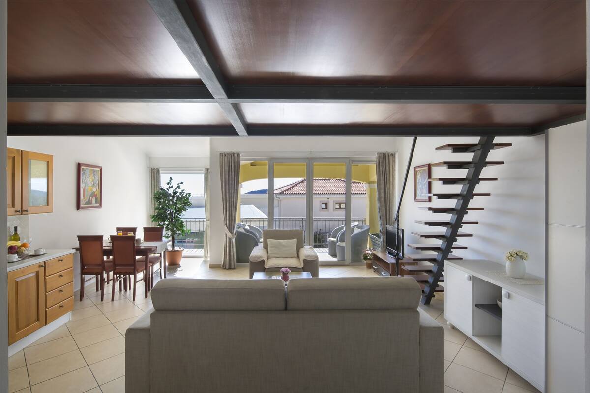 Una sala moderna con un sofá, un comedor con sillas y una mesa, una escalera y un balcón con vistas.