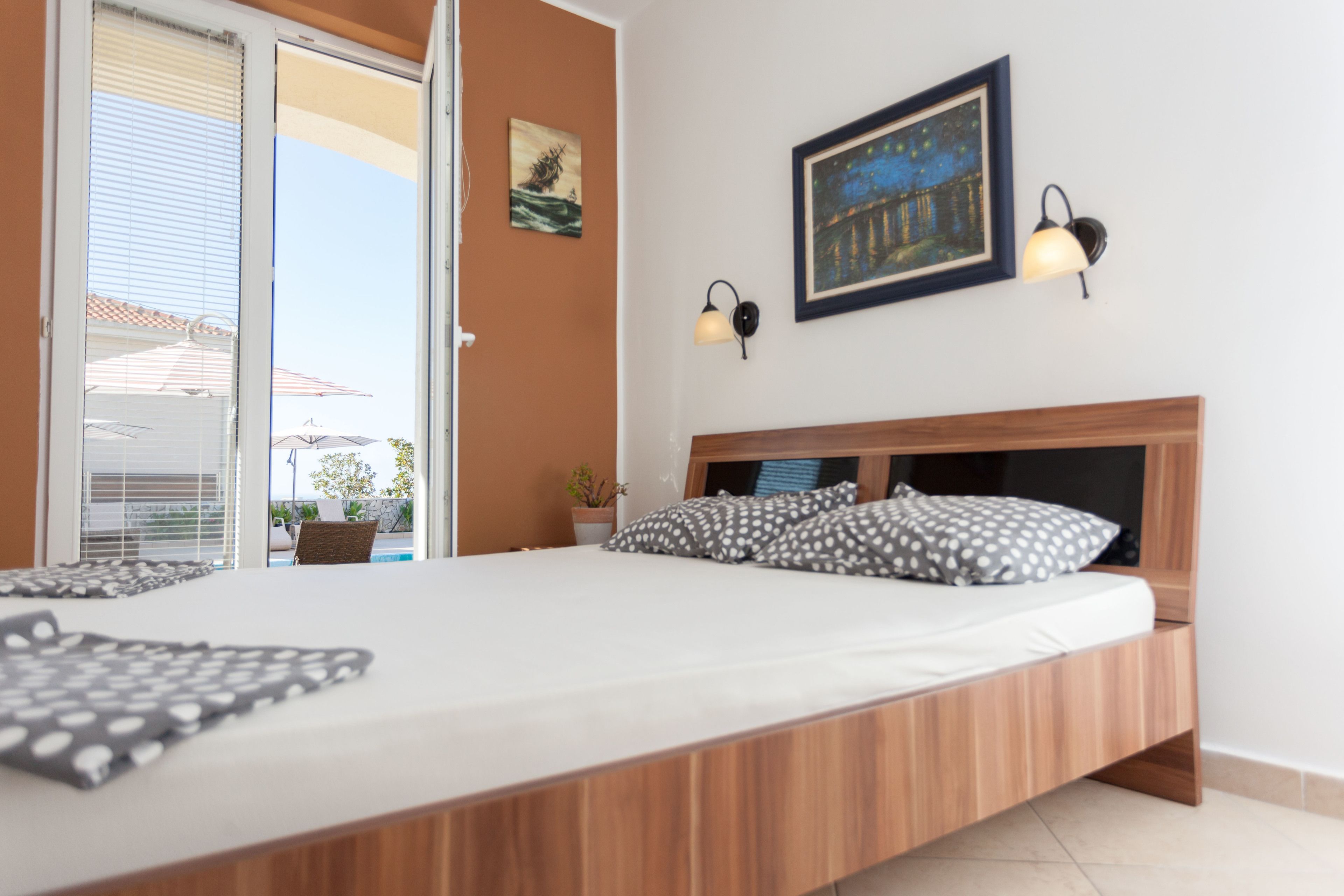 A bedroom with a wooden bed frame, a white mattress, and two pillows with a polka dot pattern. There is a window with blinds, a wall-mounted lamp, and a framed picture above the bed.