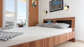 A bedroom with a wooden bed frame, a white mattress, and two pillows with a polka dot pattern. There is a window with blinds, a wall-mounted lamp, and a framed picture above the bed.