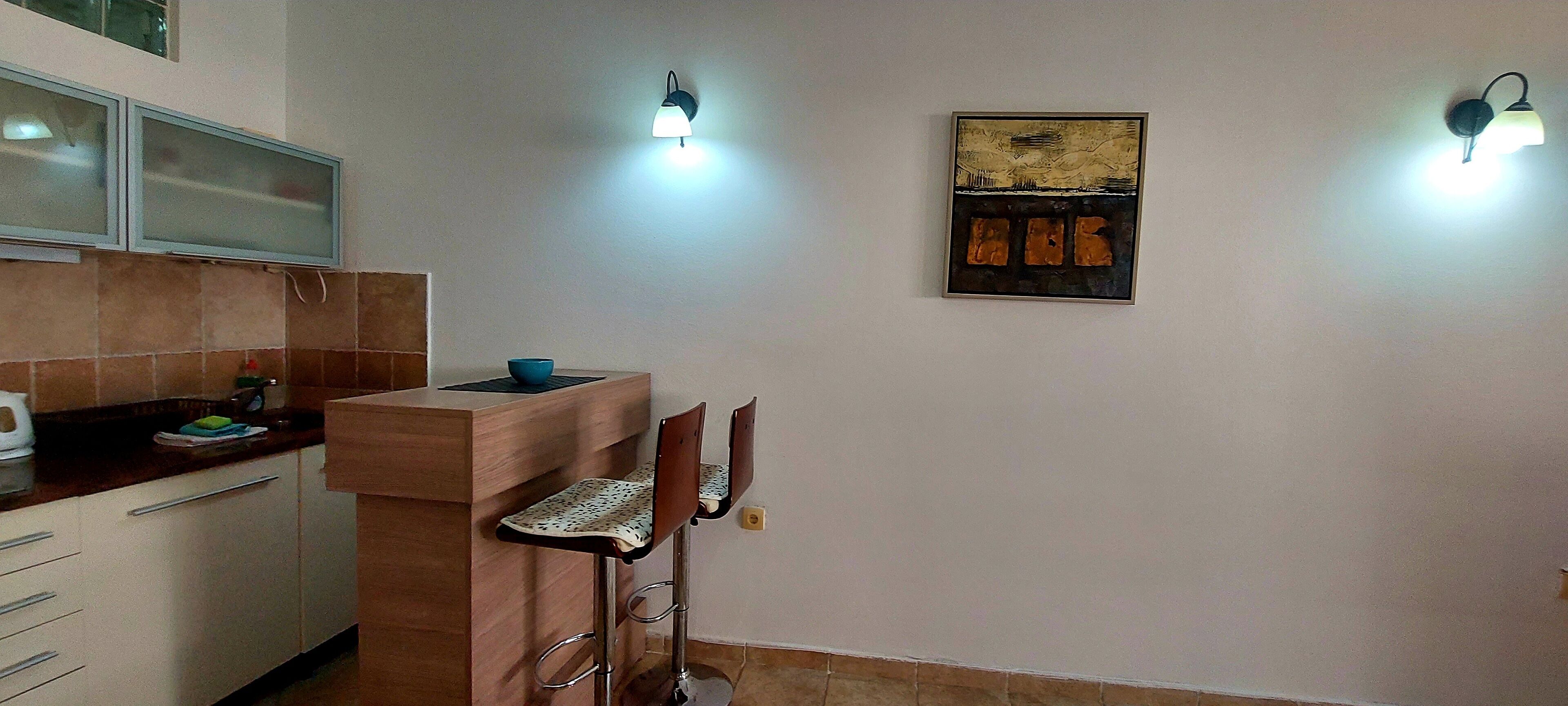 A compact kitchen with a wooden counter, two bar stools, and a wall-mounted light fixture.