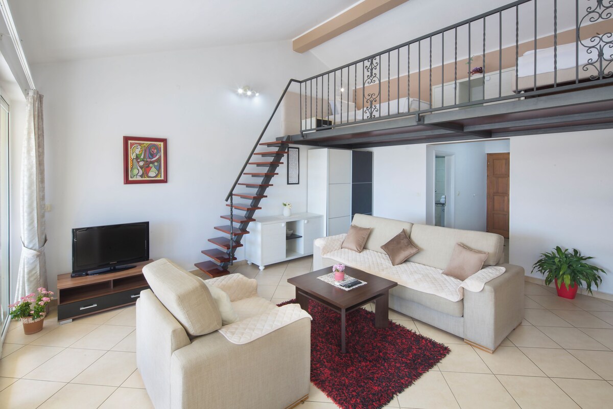 Sala de estar con sofá, mesa de centro, televisor, escalera y un balcón.