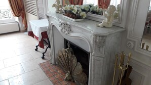 Una chimenea de mármol con intrincados relieves, una mesa blanca con mantel rojo y una figurita decorativa de un ángel encima.