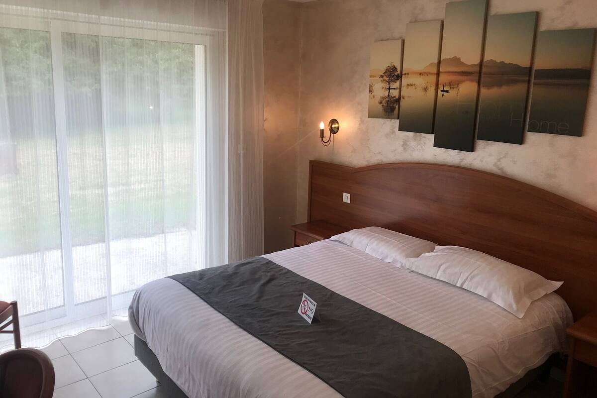 Ein Hotelzimmer mit Bett, Nachttisch, Sessel, einem Fenster mit leichtem Vorhang und Wandbildern.