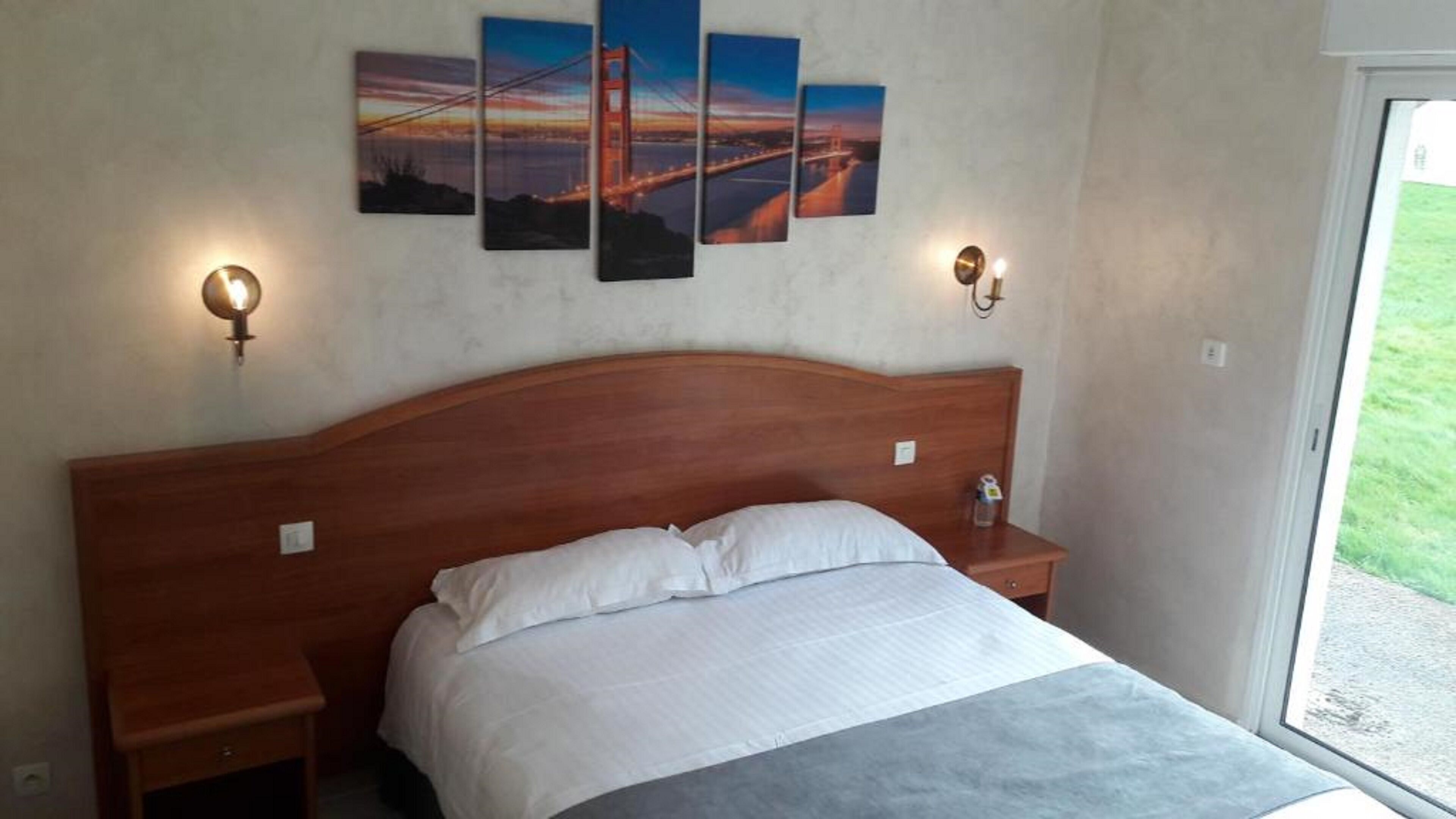 A hotel room with a wooden headboard, a bed with white linens, two bedside tables with lamps, and a large window offering a view of the outdoors.