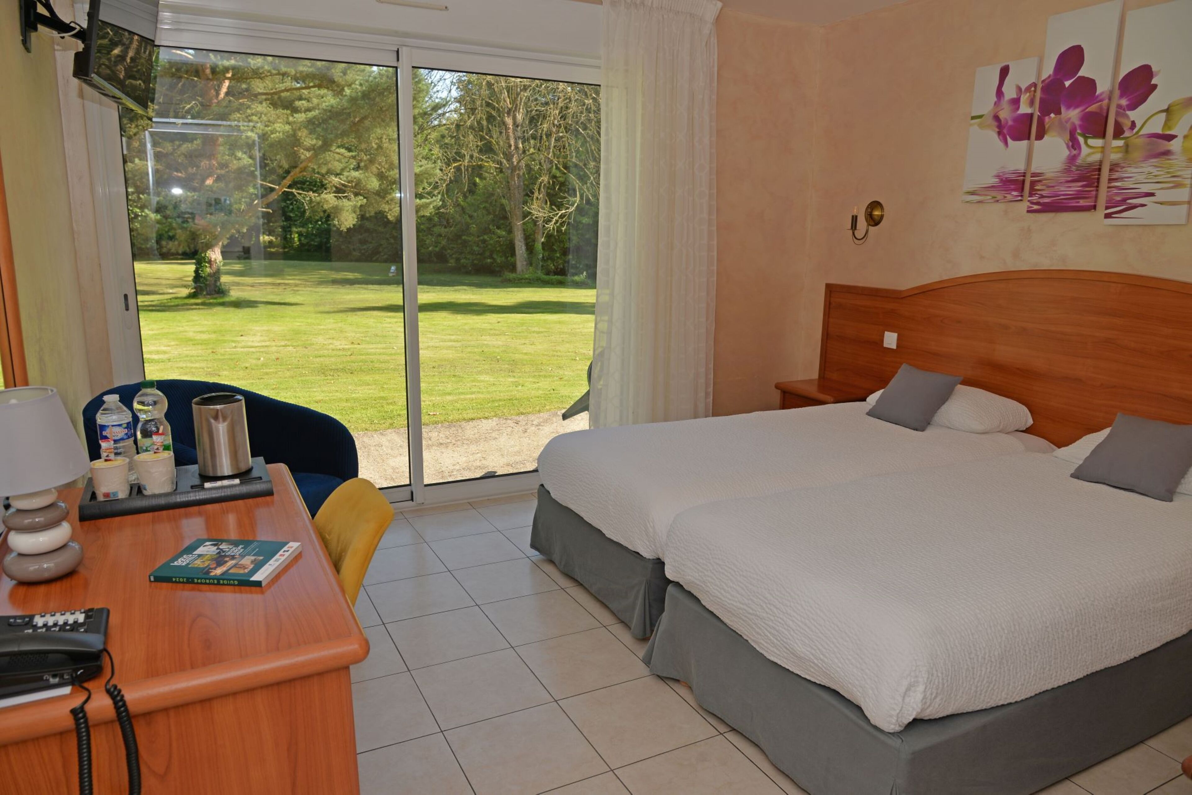 A hotel room with a large bed, a desk with a telephone, and a view of a grassy area through a sliding door.