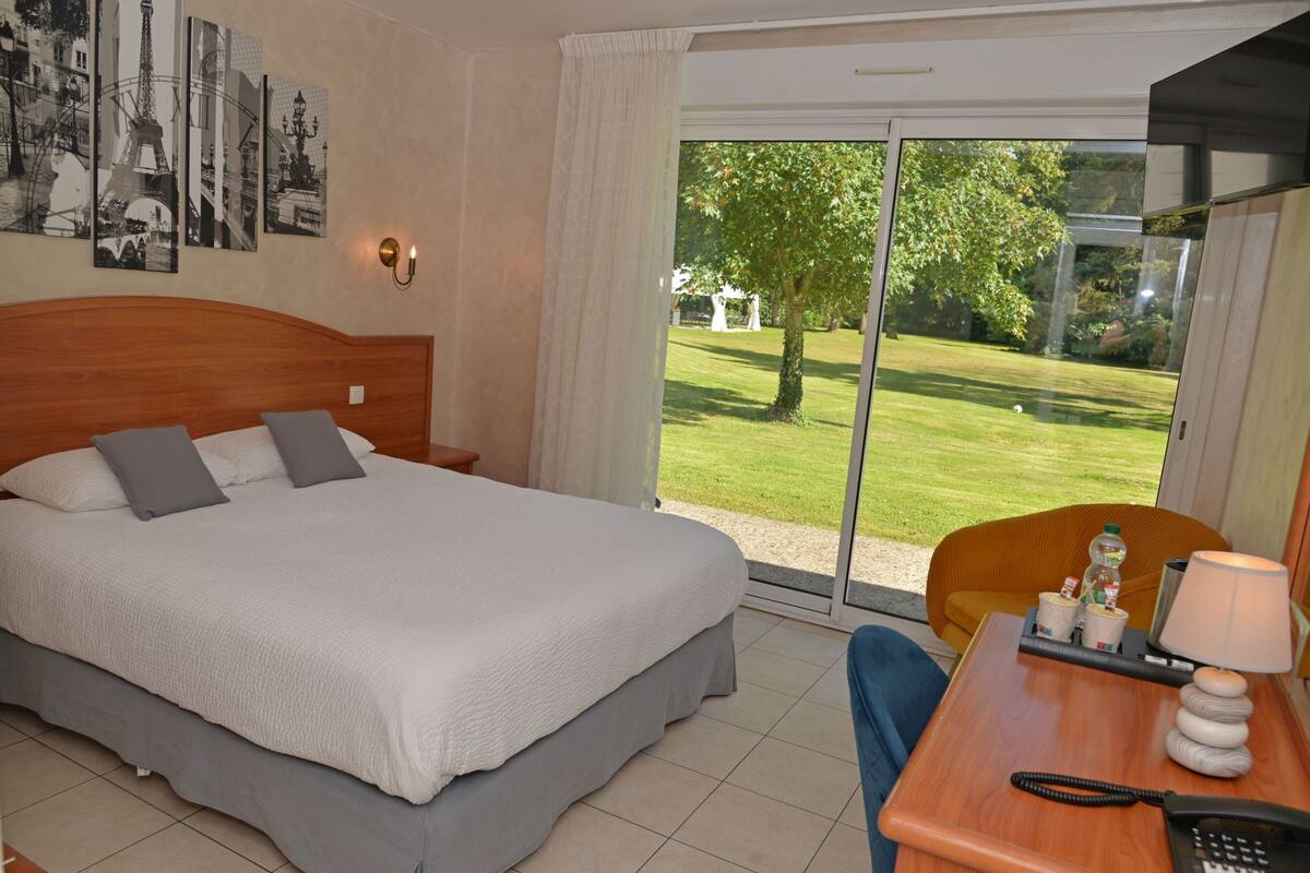 Ein Hotelzimmer mit einem großen Bett, einem Schreibtisch mit Lampe, einem Sessel und Blick auf eine grüne Rasenfläche durch eine Glasschiebetür.