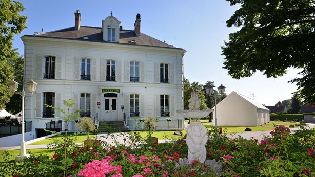 Un edificio grande y blanco con una zona de recepción, rodeado de un jardín bien cuidado con flores rosas y una estatua.