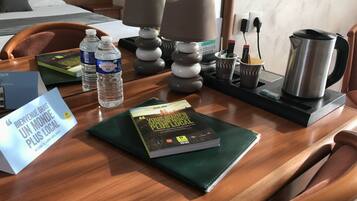 Habitación de hotel con una mesa de madera, una botella de agua, un libro, una lámpara, un hervidor y un cartel.