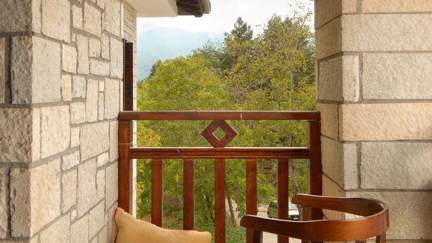 A stone-walled balcony with a wooden chair and cushions, overlooking a lush green landscape. - Mikri Arktos (Pyli)