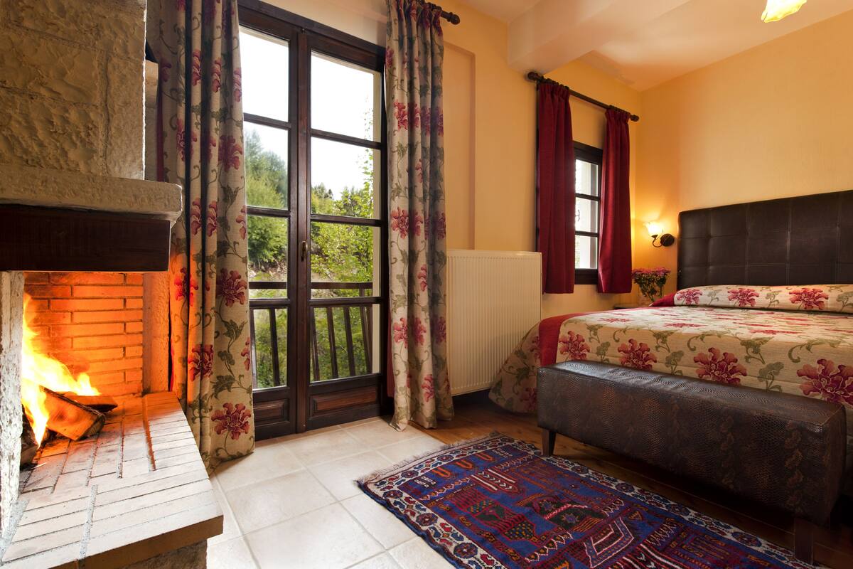 A bedroom with a fireplace, a bed with floral bedding, a bench, and a view through a large window.
