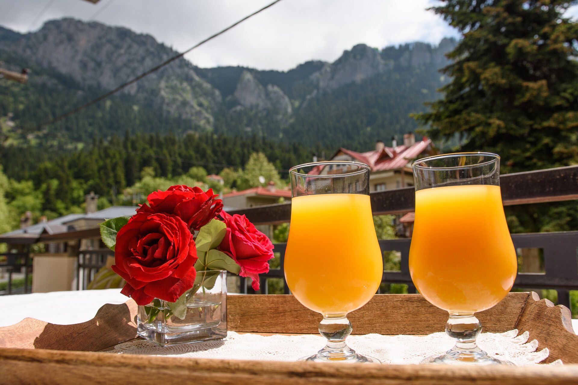 Dos vasos de jugo de naranja sobre una bandeja de madera con un florero de rosas rojas, con montañas al fondo.