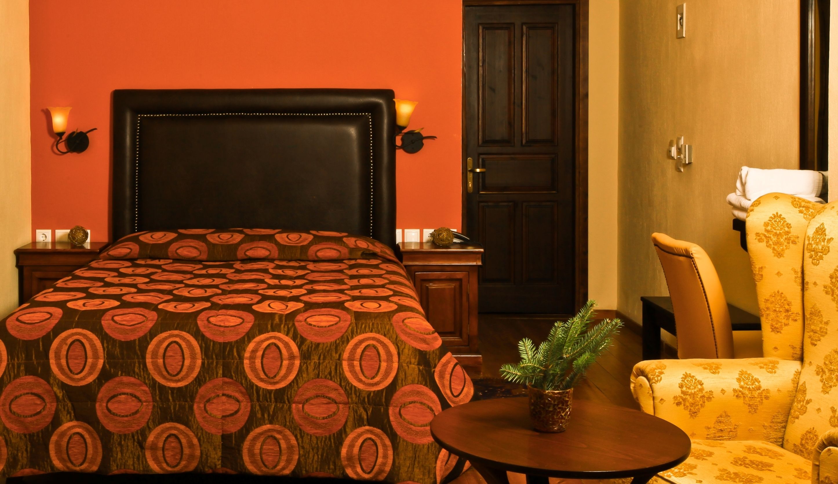 A hotel room with a large bed, a wooden nightstand, a small table with a plant, a chair, and a door.
