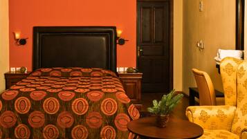 A hotel room with a large bed, a wooden nightstand, a small table with a plant, a chair, and a door.
