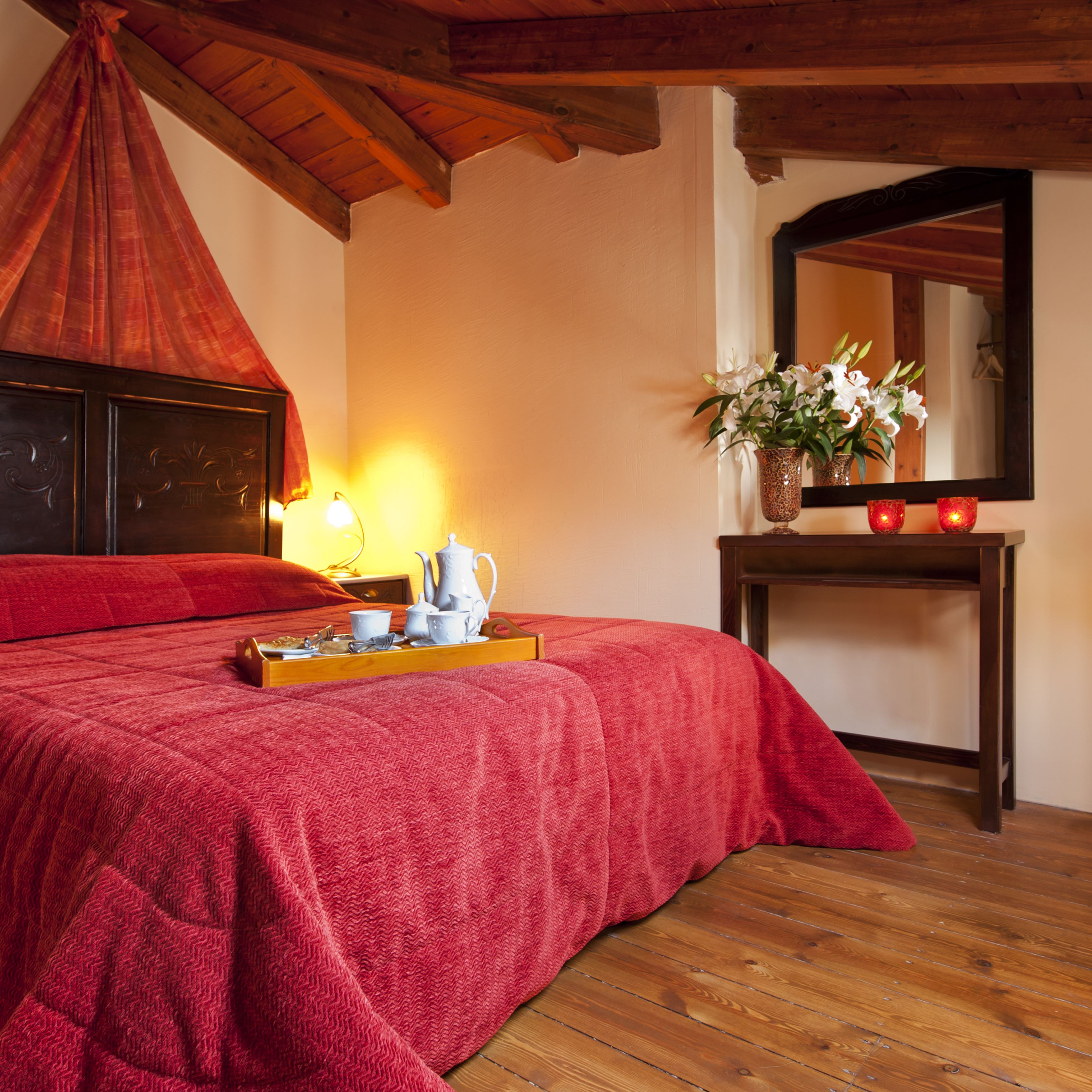Een slaapkamer met een houten plafond, een bed met een rode deken, een nachtkastje met een lamp, een vaas met bloemen, en een dienblad met theeservies.