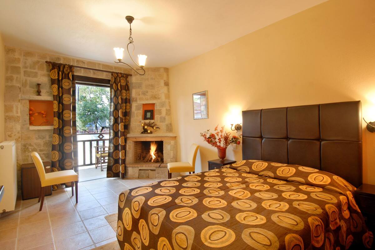 A bedroom with a fireplace, a bed with a patterned bedspread, a chair, and a nightstand with a vase of flowers.