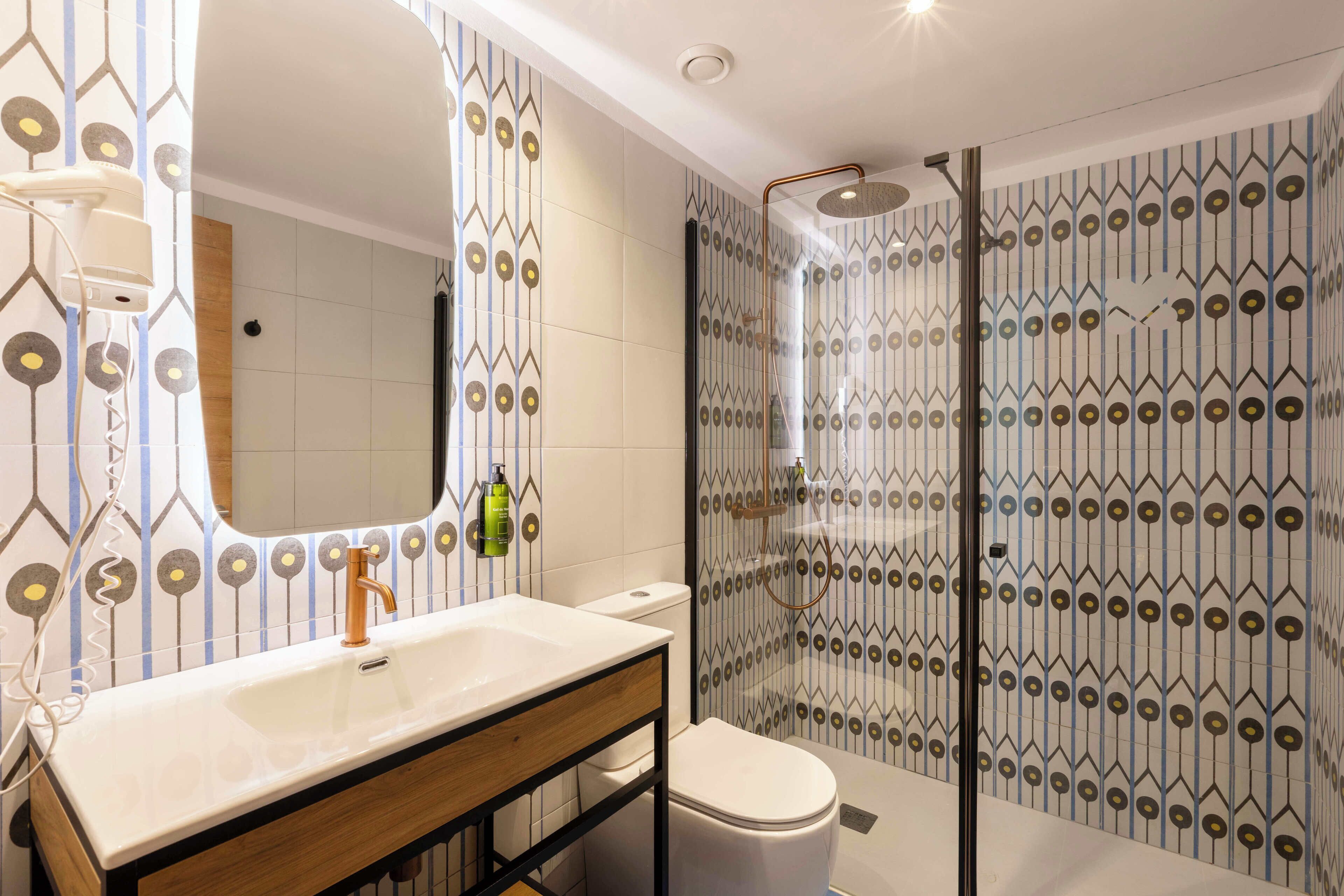 Un baño moderno con un lavabo blanco, un espejo, un inodoro y una zona de ducha con baldosas estampadas.