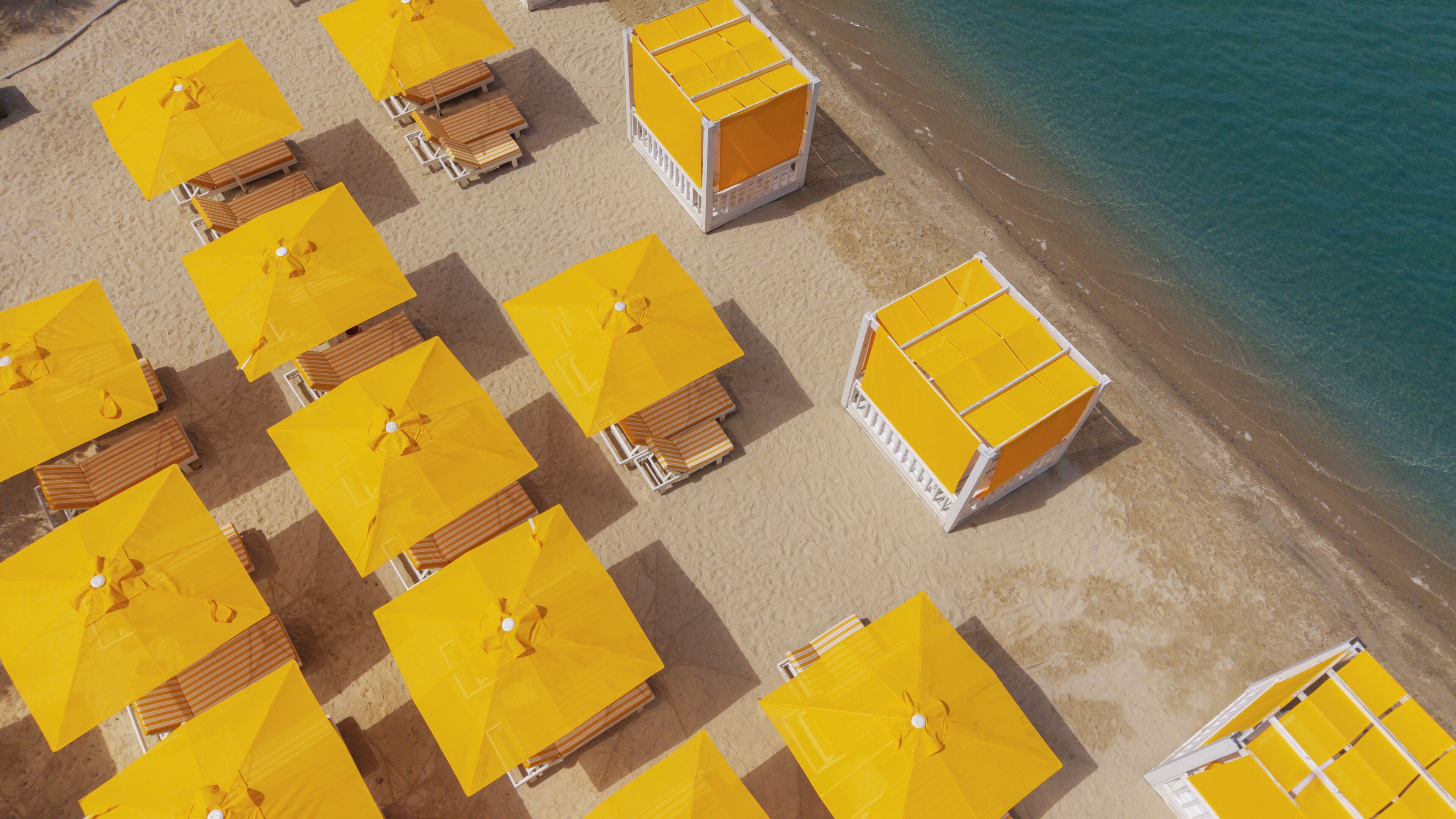 An aerial view of a beach with multiple yellow umbrellas and wooden lounge chairs arranged in rows.