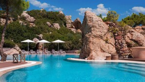 Piscina con sillas de descanso y sombrillas, rodeada de terreno rocoso y árboles.