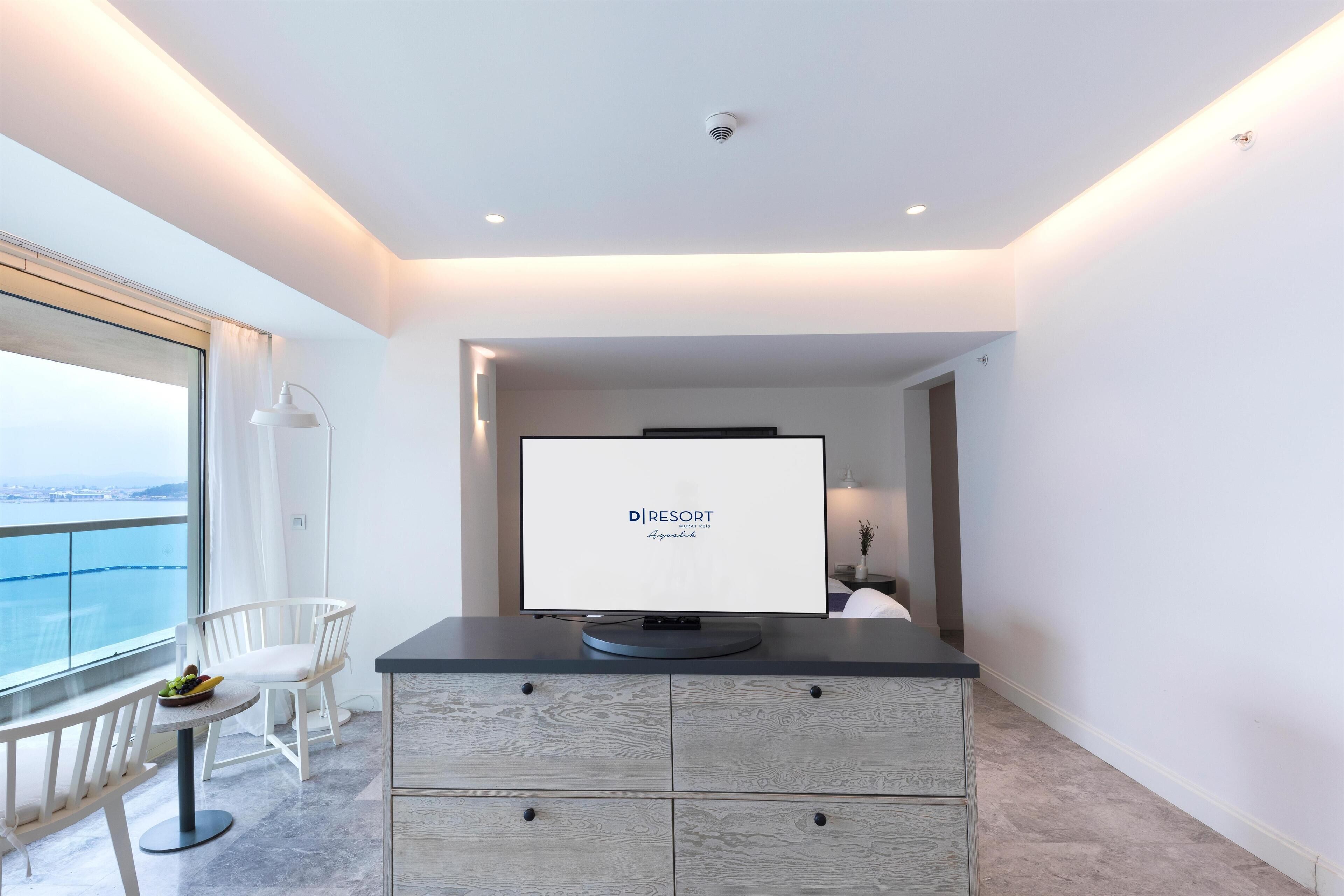 Habitación de hotel moderna con un ventanal grande que ofrece vistas a la ciudad, un televisor de pantalla plana sobre un mueble oscuro, una silla blanca y una mesa pequeña con fruta.