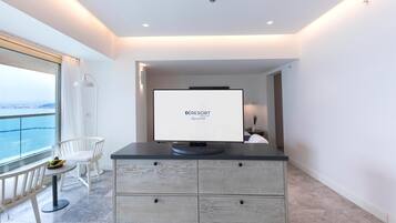 Habitación de hotel moderna con un ventanal grande que ofrece vistas a la ciudad, un televisor de pantalla plana sobre un mueble oscuro, una silla blanca y una mesa pequeña con fruta.