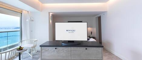 A modern hotel room with a large window offering a city view, a flat-screen TV on a dark console, a white chair, and a small table with fruit.