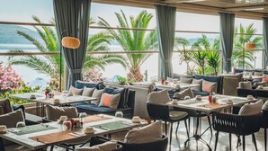 Un restaurante con mesas y sillas dispuestas para comer, ventanas amplias con cortinas y vistas al mar y palmeras.