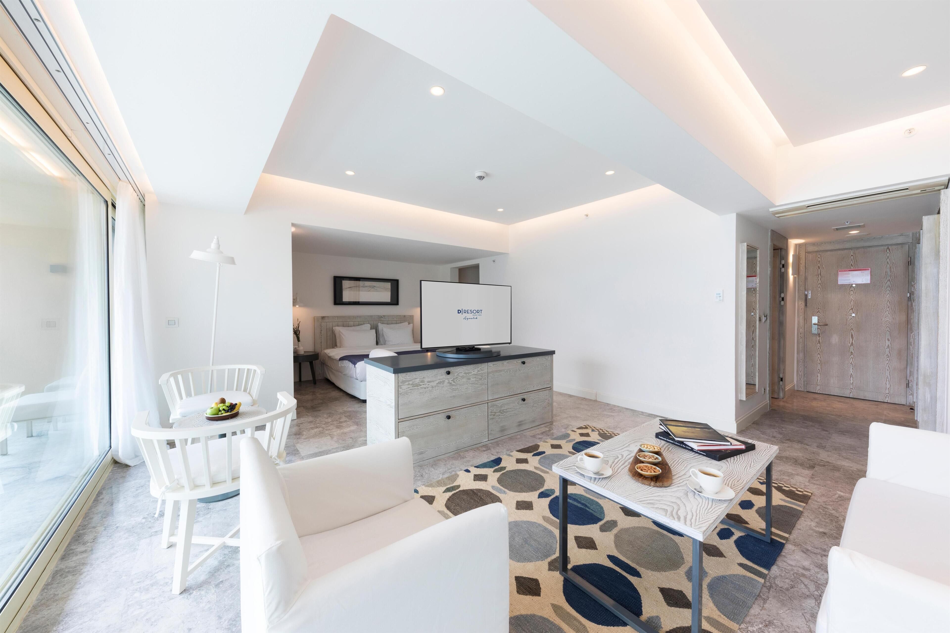 Una habitación de hotel moderna con zona de comedor, sofá, mesa de centro y televisión.