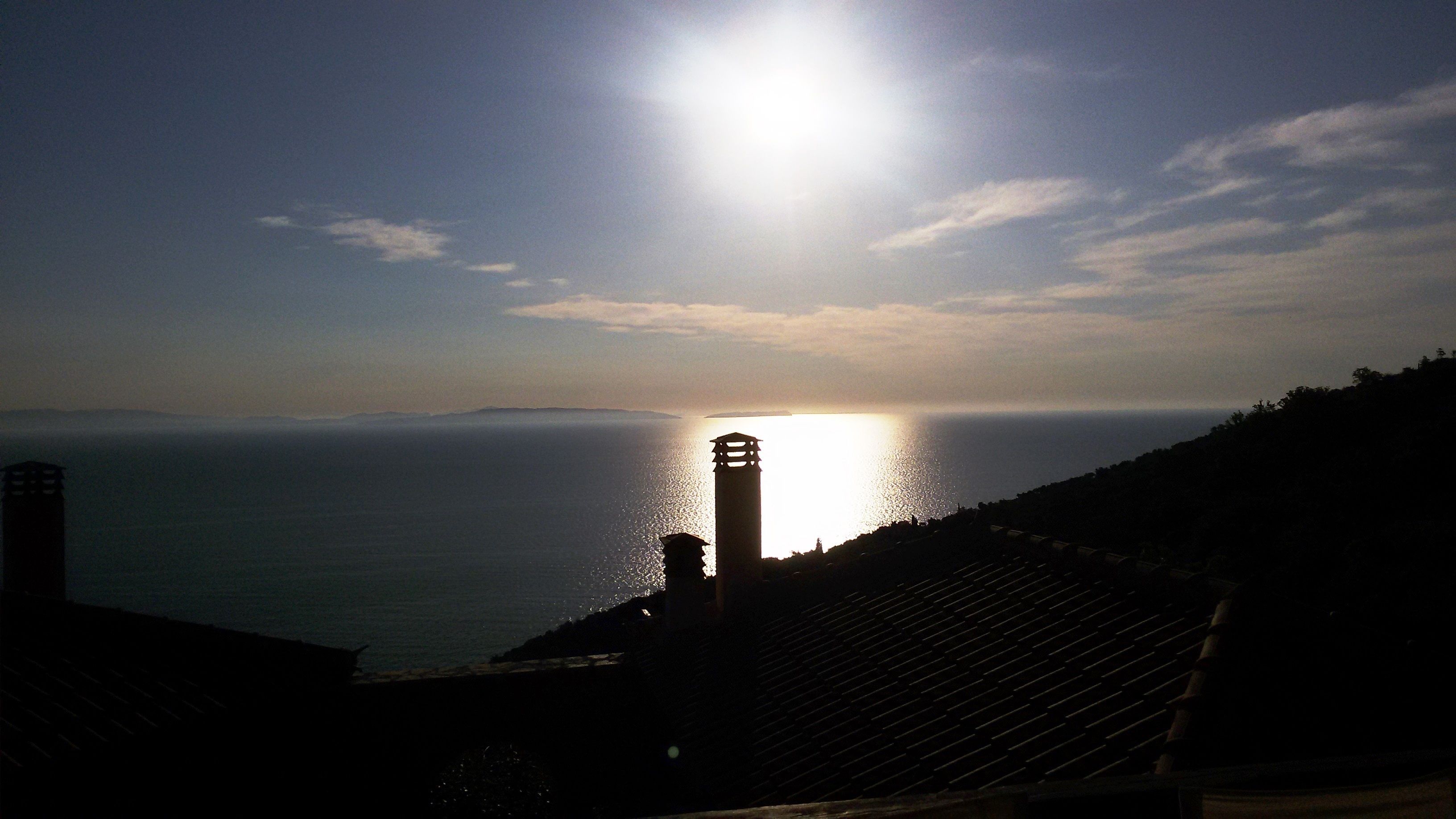 Vista costera con la silueta de un faro, cielo despejado y el sol ocultándose o saliendo sobre el agua.
