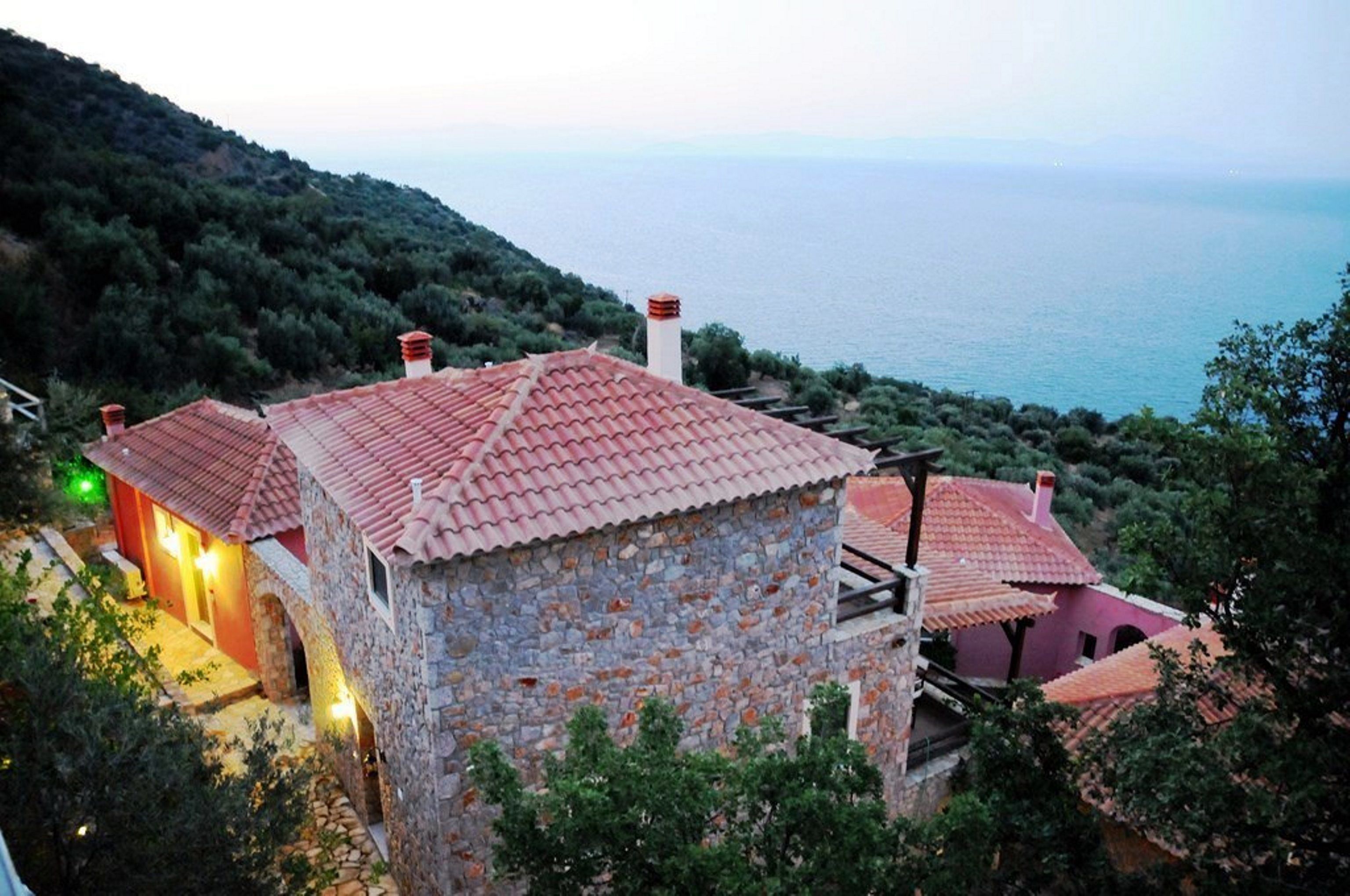 A stone building with a red-tiled roof, situated on a hillside overlooking a body of water.