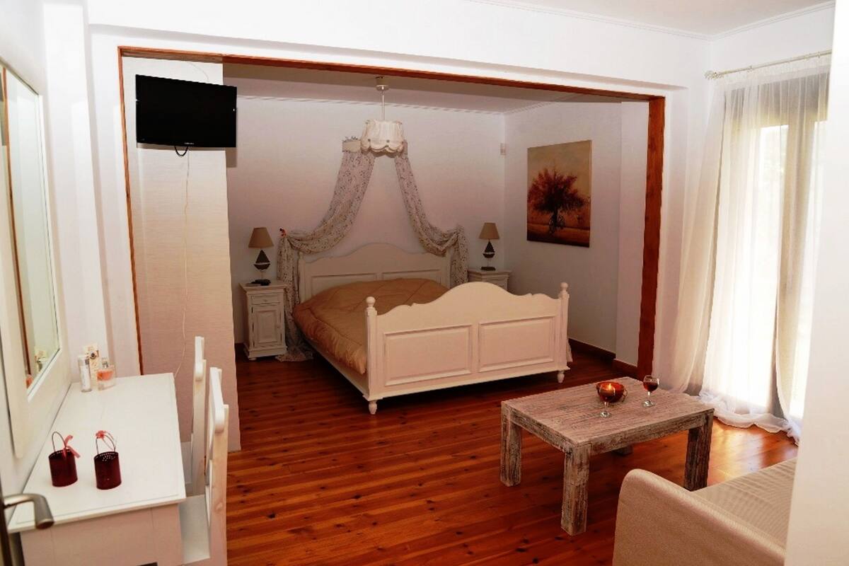 Ein Schlafzimmer mit Holzboden, einem weißen Bett mit Baldachin, einem Nachttisch mit Lampe, einem kleinen Holztisch mit Kerze und Gläsern sowie einem Gemälde an der Wand.