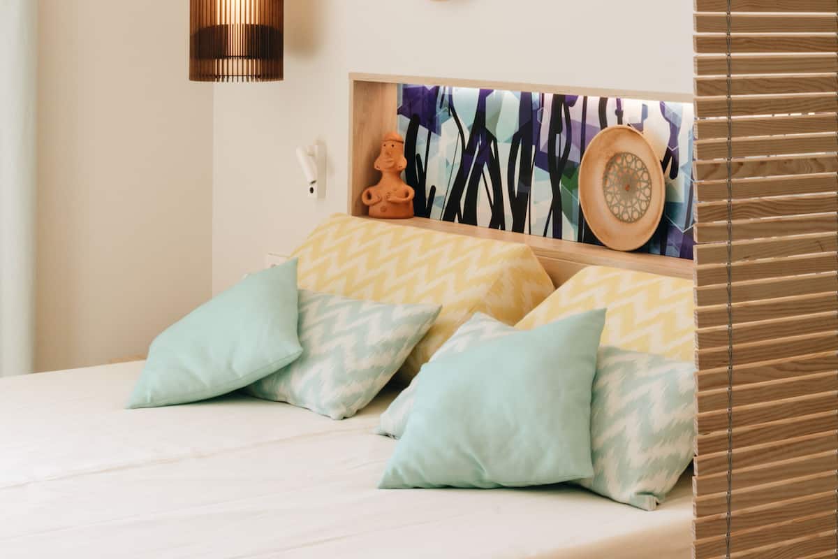 Un lit avec des oreillers à motifs verts clairs et jaunes, une tête de lit en bois et une étagère murale agrémentée d’objets décoratifs.