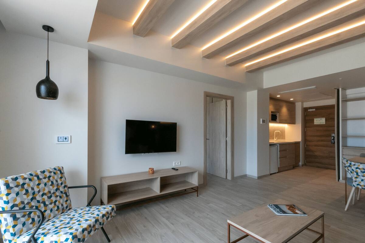 Un salon moderne avec un téléviseur à écran plat, un canapé à motifs, une table basse en bois et un coin cuisine à l’arrière-plan.