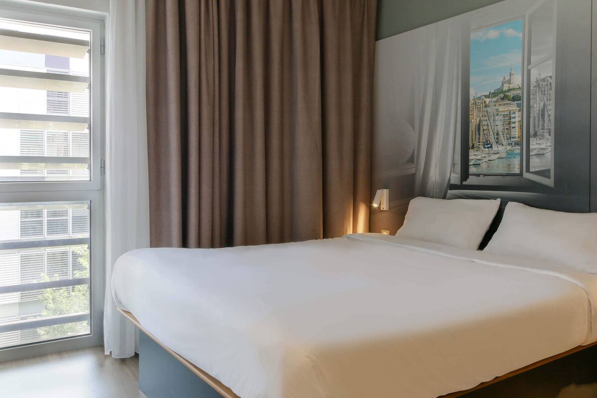 A neatly made bed in a hotel room with a large window, curtains, and a painting of a cityscape.