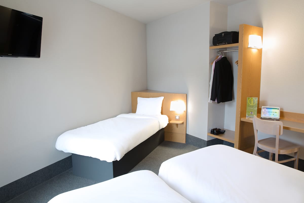 A hotel room with two single beds, a desk with a chair, a TV mounted on the wall, and a coat rack with a jacket.