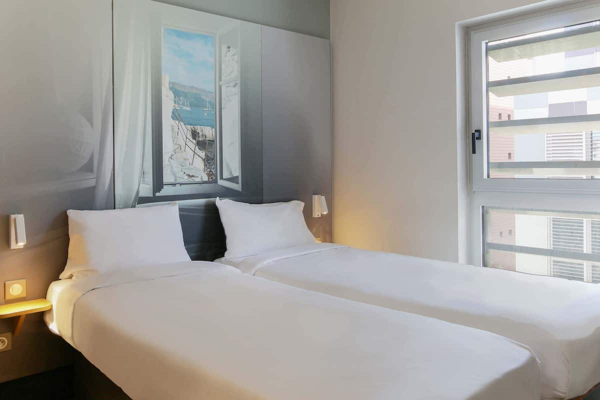 A hotel room with a bed, a nightstand, a window with blinds, and a painting on the wall.